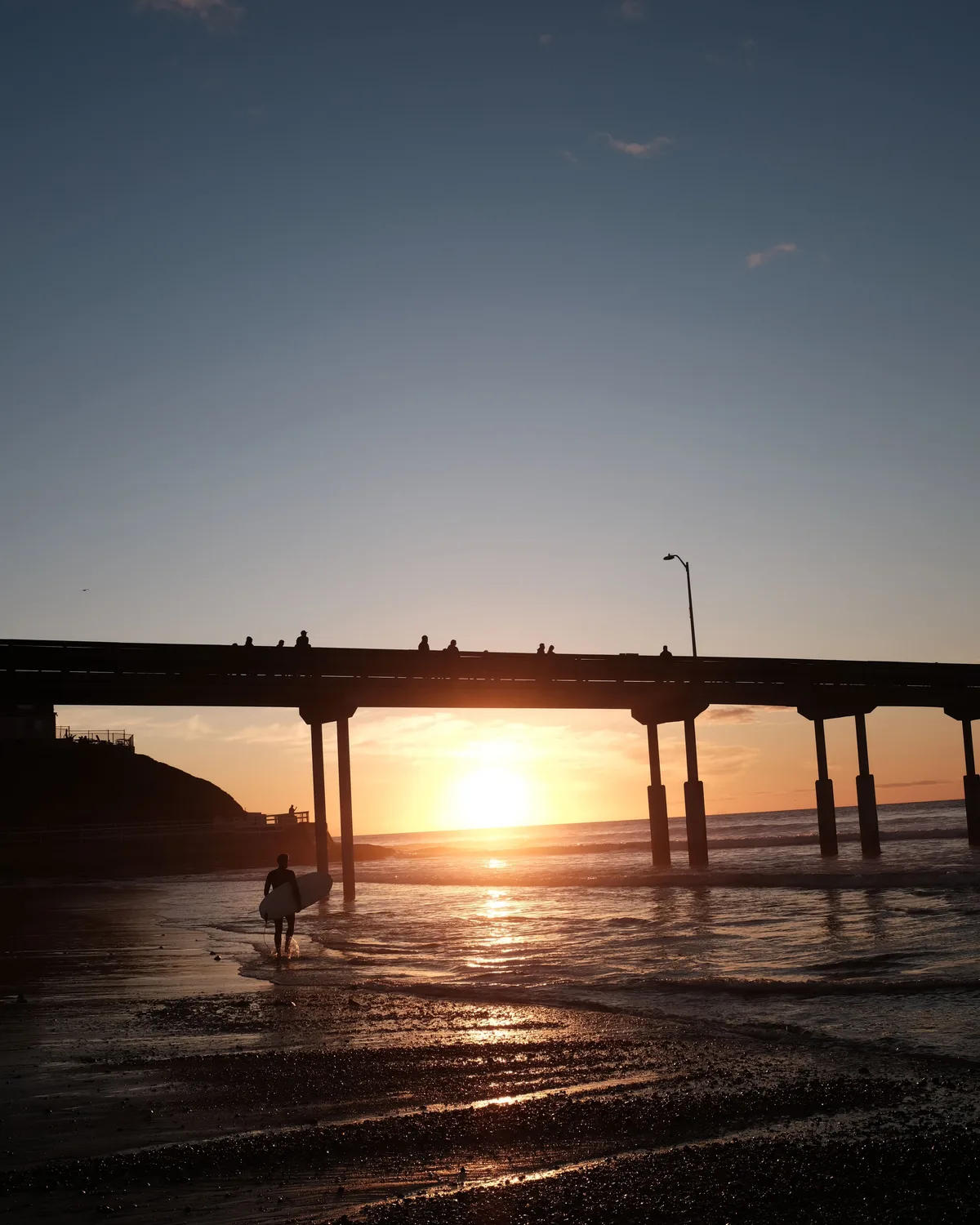 A surfer walking beneath Ocean Beach Pier at sunset